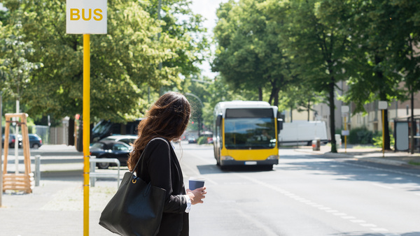 Eine Frau wartet an einer Bushaltestelle auf den herbeifahrenden gelben Bus und hat einen Kaffeebecher in der Hand.