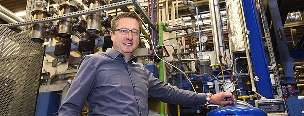 Ein farbiges Foto zeigt Christian Schwierz, den Leiter der Härterei von IMS Gear, in der industriellen Anlage. Er steht vor Maschinen und Rohrleitungen und hält die Hand auf einen blauen Druckbehälter mit Manometer.