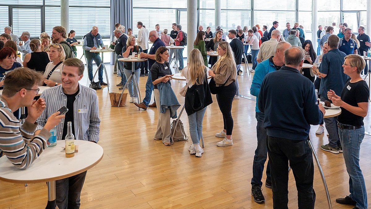 Farbige Fotografie: Viele Menschen tauschen sich bei einem Stehempfang aus. Getränke und Teller stehen auf hohen Tischen.