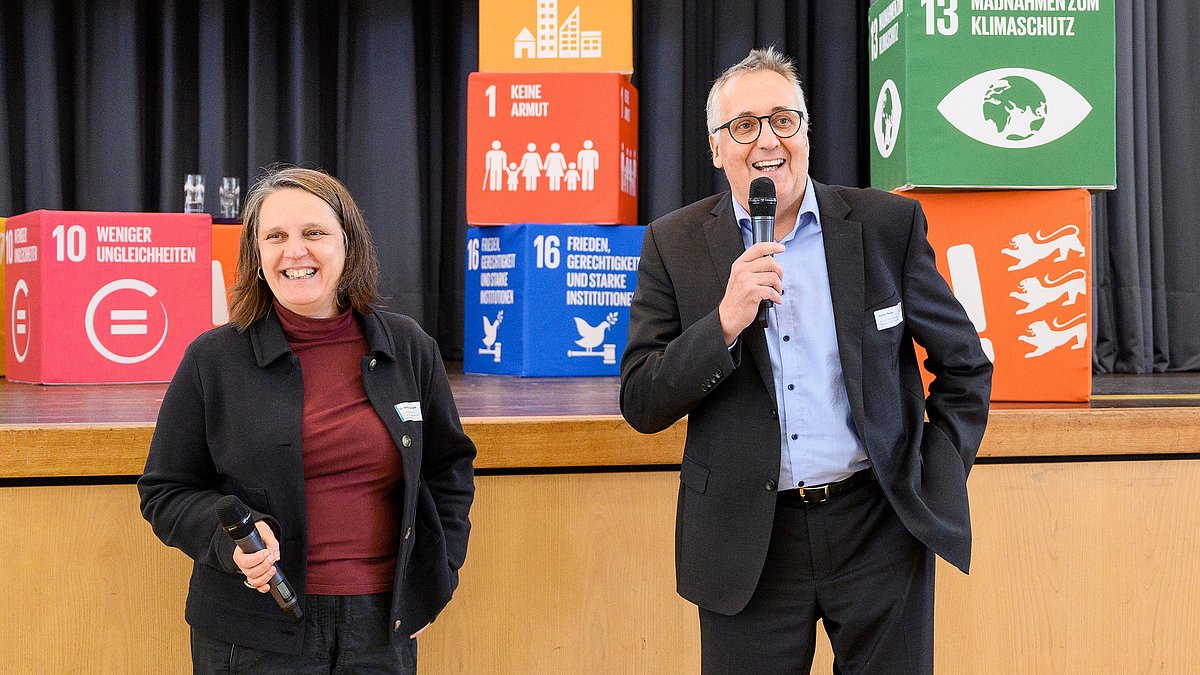 Frau Dengler und Herr Beule stehen mit Mikrophonen in der Hand vor der Bühne und unterhalten sich freudig.