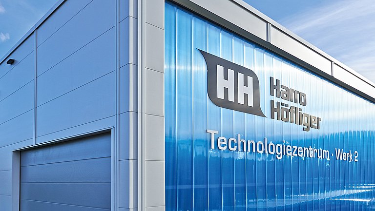 Das Bild zeigt ein Teil der Gebäudes von Harro Höfliger mit einer Glasfront und dem Unternehmensnamen darauf.