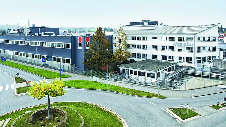 Große dunkelgraue Halle neben einem weißen Bürogebäude. Davor eine Straße mit einem Kreisverkehr.