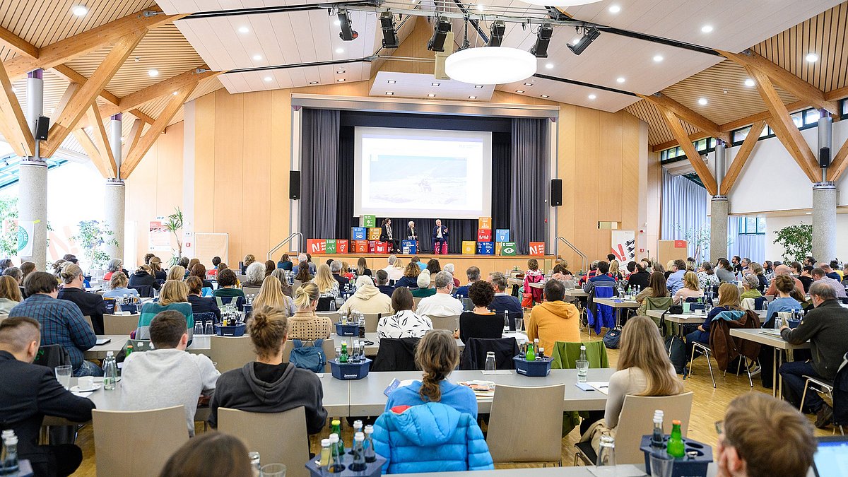 Das Bild zeigt eine Veranstaltungshalle mit Blick auf die Bühne und vielen Personen, die an Tischen sitzen und zur Bühne schauen.