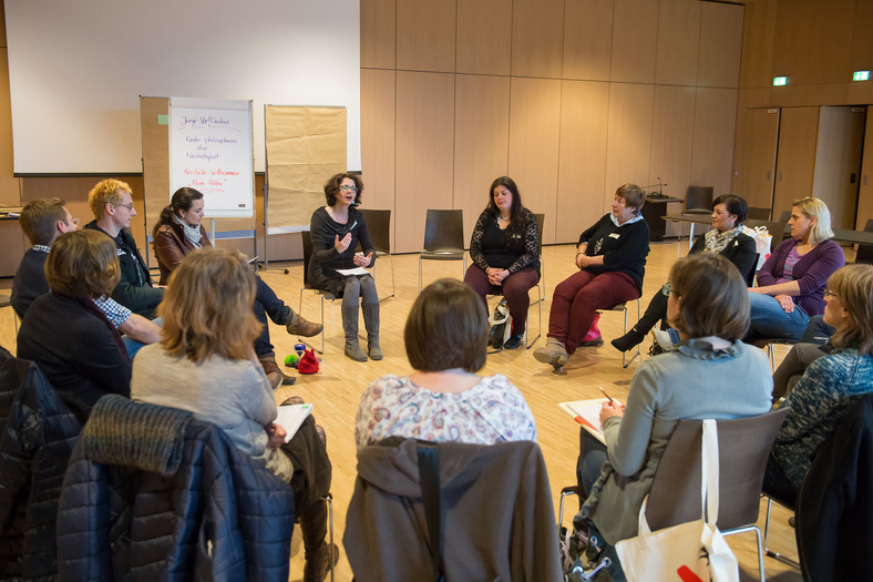 Ein Saal mit Stuhlkreis. Dreizehn Personen sitzen, eine Frau spricht zu ihnen. Im Hintergrund stehen Flipcharts, dahinter eine Leinwand.