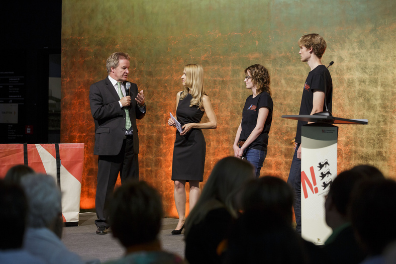 Nachhaltigkeitstage 2014 Umweltminister Untersteller auf der Bühne mit zwei Jugendbeiräten der Jugendinitiative der Nachhaltigkeitsstrategie. Er hält das Mikrofon und blickt die Moderatorin an.