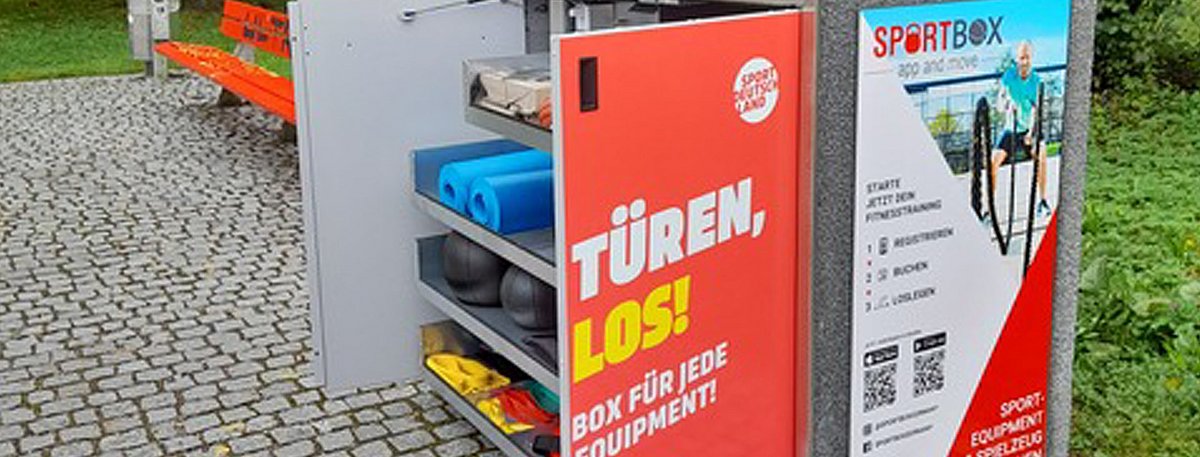 Am Rande einer Parkfläche steht eine große Box mit geöffneten Türen, die einen Blick auf verschiedene Sportgeräte freigeben.