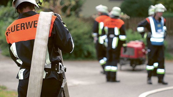 Dargestellt sind Feuerwehrleute im Einsatz. Einer der Feuerwehrmänner trägt einen Schlauch über der Schulter.