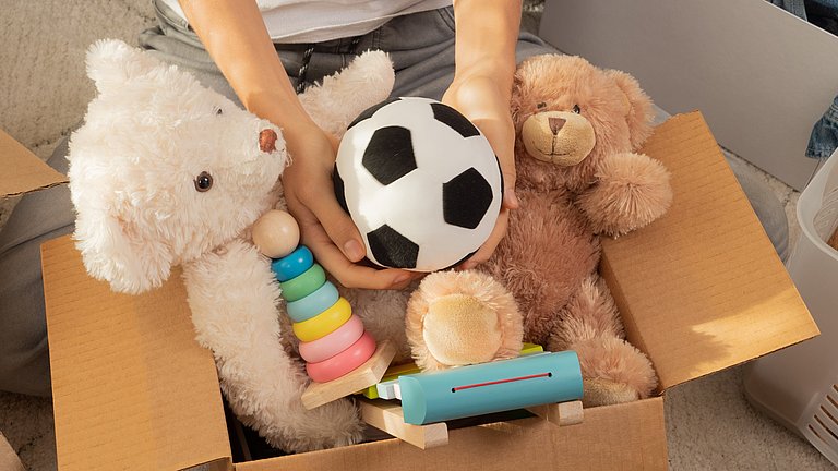 In eine offene Pappkiste legt ein Kind zu vielen Spielsachen einen Ball.