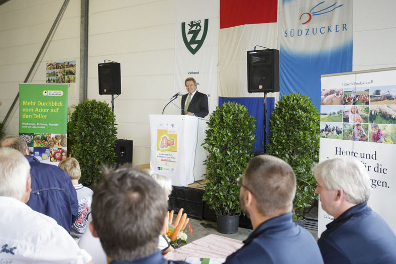 Minister Untersteller in einer spricht vor Publikum. Im Hintergrund Banner mit Südzucker-Logo und "Gläserne Produktion". 