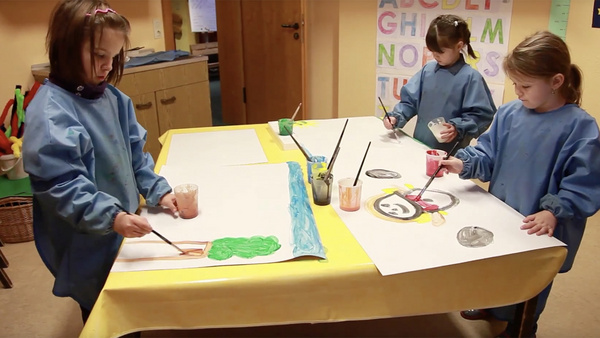 Drei Kindergartenkinder in Malkitteln malen mit Wasserfarben an einem Tisch.