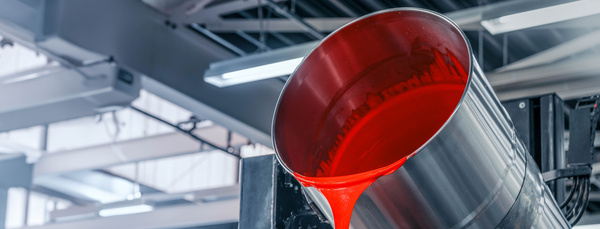 In einer hellen Produktionshalle fließt aus einem großen Metallfass rote Farbe in den Container einer Abfüllanlage.