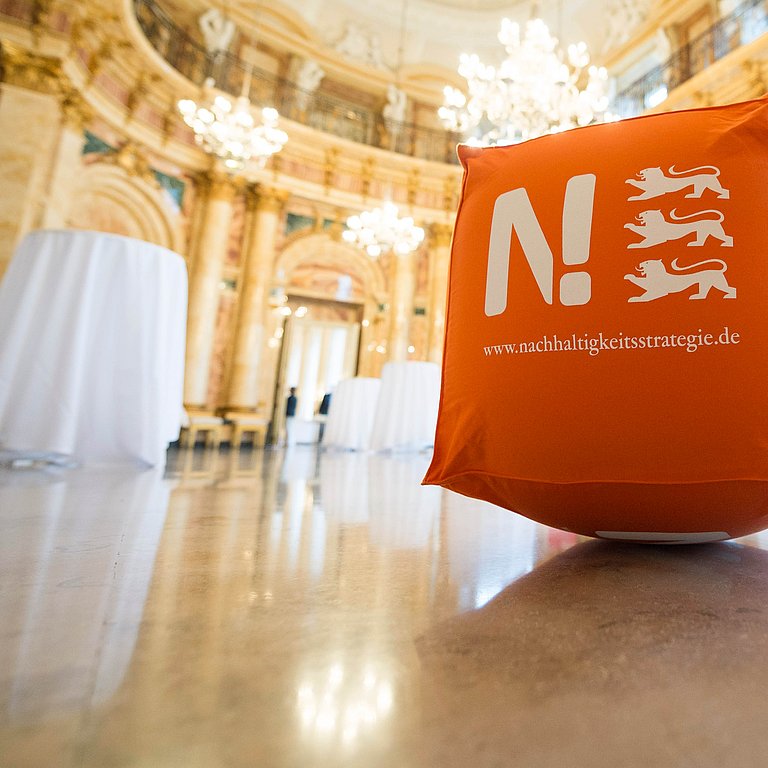In einem prunkvollen Saal mit Kronleuchtern steht ein orangener Würfel mit dem Logo der Nachhaltigkeitsstrategie.