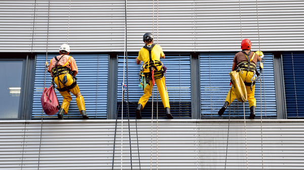 Dargestellt sind drei Fensterputzer, die frei hängend an einer Bürofassade die Fenster reinigen.