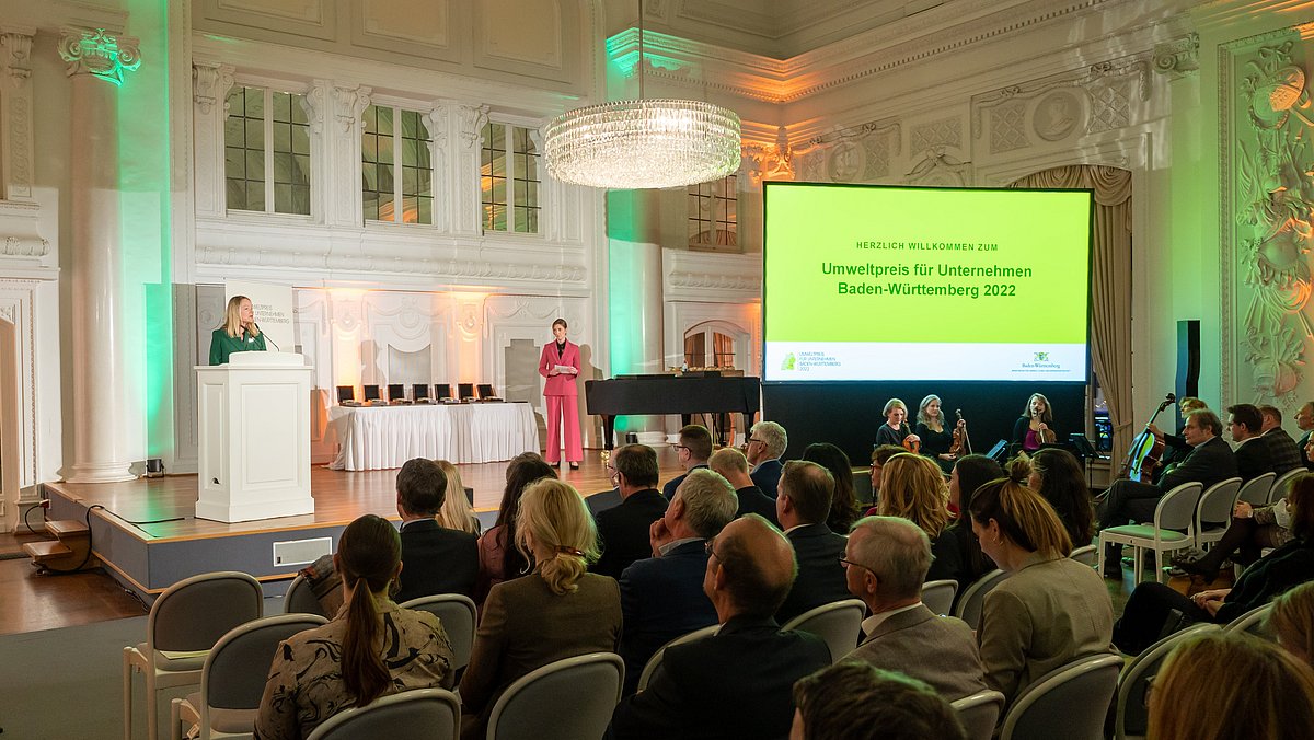 Umweltministerin Thekla Walker steht am Rednerpult auf der Bühne, auf der eine weitere Frau sowie eine Leinwand mit der Aufschrift "Umweltpreis für Unternehmen Baden-Württemberg 2022" stehen. Vor der Bühne sitzen mehrere Menschen mit Blick zur Bühne.