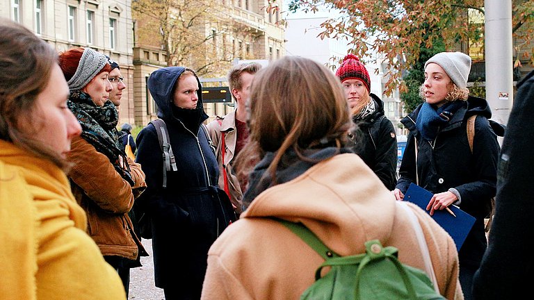 Eine Gruppe von über 8 Frauen und Männer erhalten im Rahmen einer Stadtführung Informationen über Stuttgart.