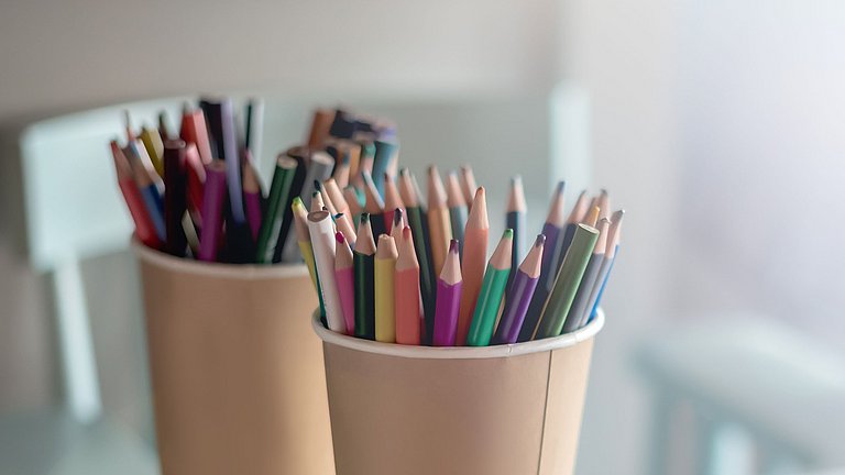 Zwei Pappbecher, die mit bunten Stiften gefüllt auf einem Tisch stehen. 