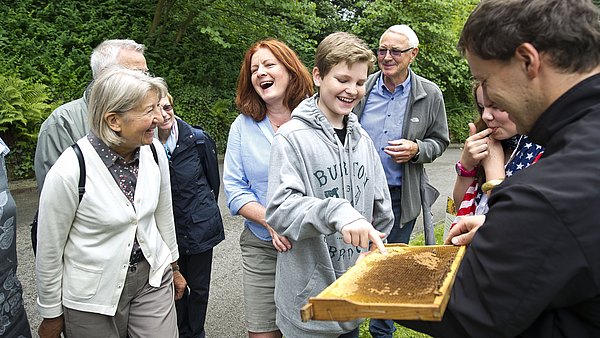 Ein Mann hält Bienenwaben aus dem Bienenstock. Ein Junge fasst sie an, lächelt. Menschen um ihn herum lachen.