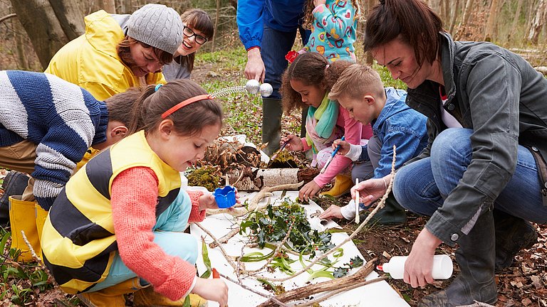 Kinder und mehrere Erwachsene breiten Blätter und anderes Naturmaterial auf einer weißen Fläche aus. Ein Mädchen pinselt.