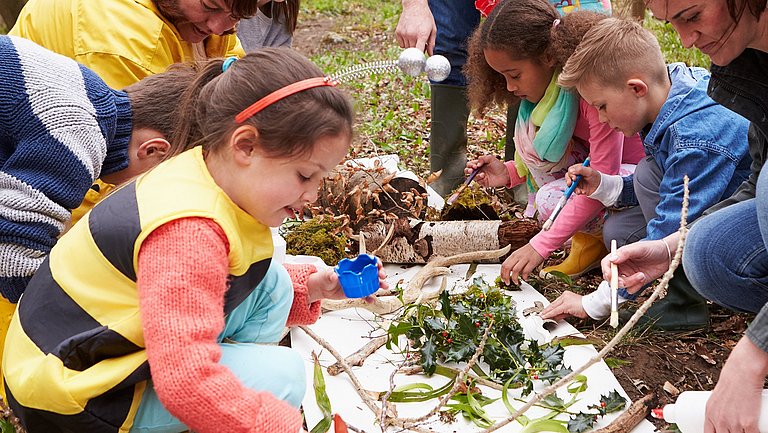 Kinder und mehrere Erwachsene breiten Blätter und anderes Naturmaterial auf einer weißen Fläche aus. Ein Mädchen pinselt.