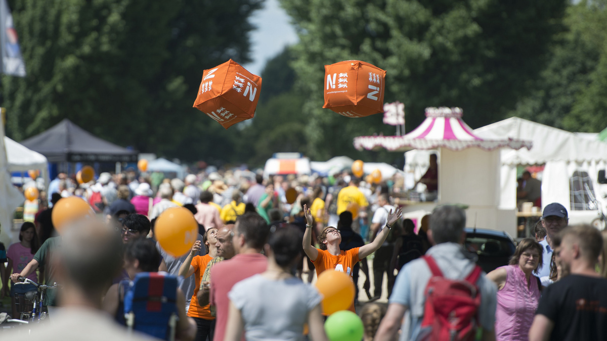 Vielen Menschen tummeln sich bei einer Veranstaltung der Nachhaltigkeitsstrategie. Zwei orangefarbene Würfel fliegen schweben über den Besuchern.
