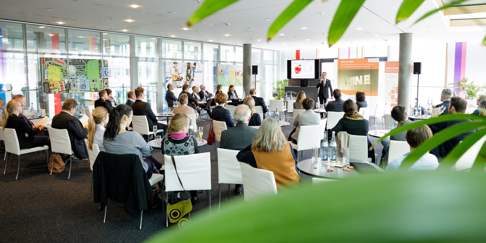 Interessierte und Unternehmen sitzen während WIN Forum an Tischen und schenken einem Vortrag ihre Aufmerksamkeit.