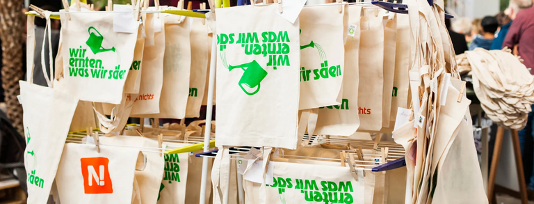 Siebdruckwerkstatt auf einer Messe: Jutetüten mit dem Aufdruck "Wir ernten was wir säen" sind aufgehängt zum Trocknen