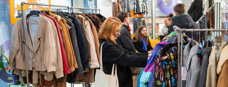 Konsum & Lifestyle Junge Frau schaut sich Kleidung an. Im Hintergrund gibt es einen Workshop zum Thema Nachhaltiger Konsum.